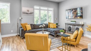 A modern living room with a sectional sofa, a yellow armchair, and a coffee table. There are two windows with blinds, a flat-screen TV, and a small table with decorative items. - Nashville Nest - Rooftop Patio - Mins To Broadway (Nashville)