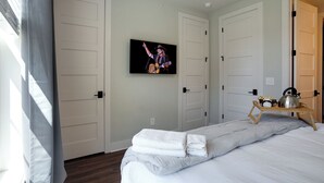 Une chambre d’hôtel avec un lit, un téléviseur fixé au mur et une étagère en bois sur laquelle se trouvent une bouilloire et des fleurs.