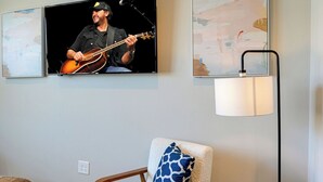 Un salon avec un téléviseur diffusant un programme mettant en scène un musicien, un lampadaire moderne, un fauteuil rembourré et un tapis à motifs.