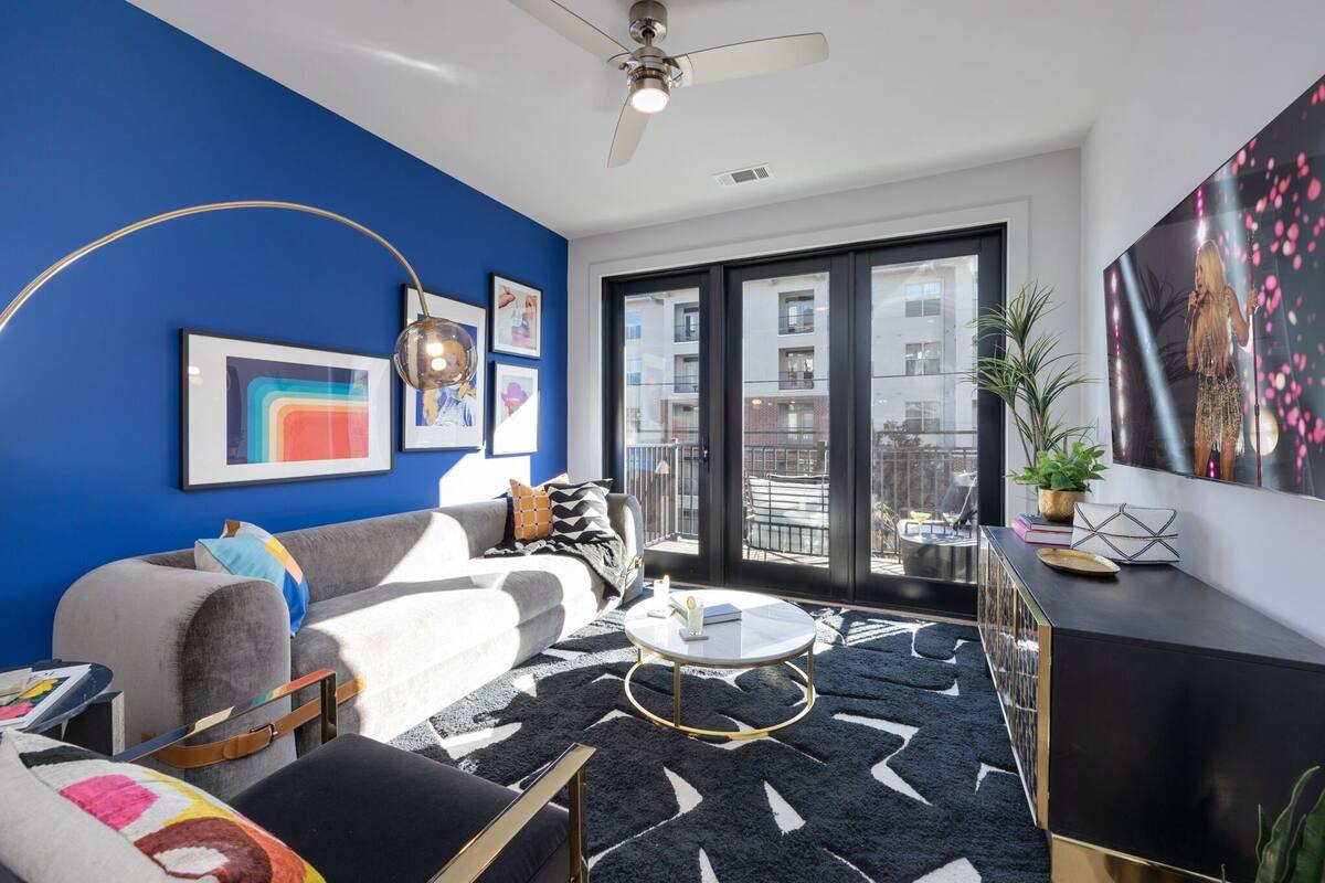 A modern living room with a blue accent wall, a ceiling fan, a large window with a view of buildings, a black and white rug, and a black and gold console table.