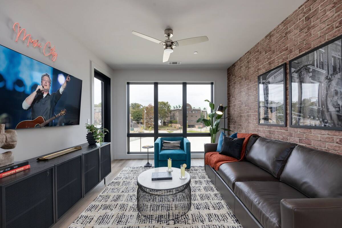 A modern living room with a large TV, a ceiling fan, a brick wall, and a large window.
