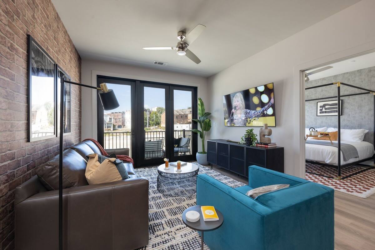 A modern living room with a sofa, a coffee table, and a blue armchair. There is a balcony with a view of buildings.