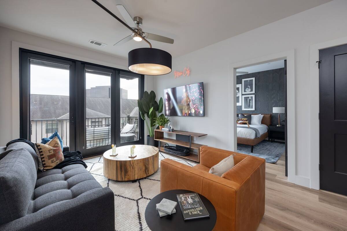 A modern living room with a grey sofa, a wooden coffee table, and a black side table. There is a large window with a view of buildings, a ceiling fan, and a black pendant light.
