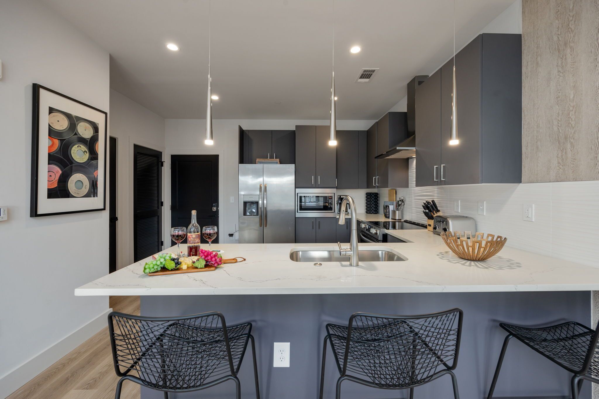 A modern kitchen with a central island, stainless steel appliances, pendant lighting, and black chairs.