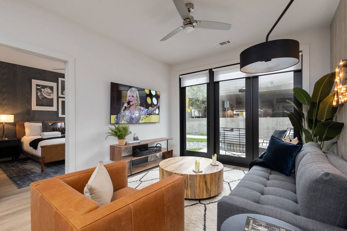 A modern living room with a sofa, armchair, and coffee table, a TV mounted on the wall, and a large window with a view of the outdoors.