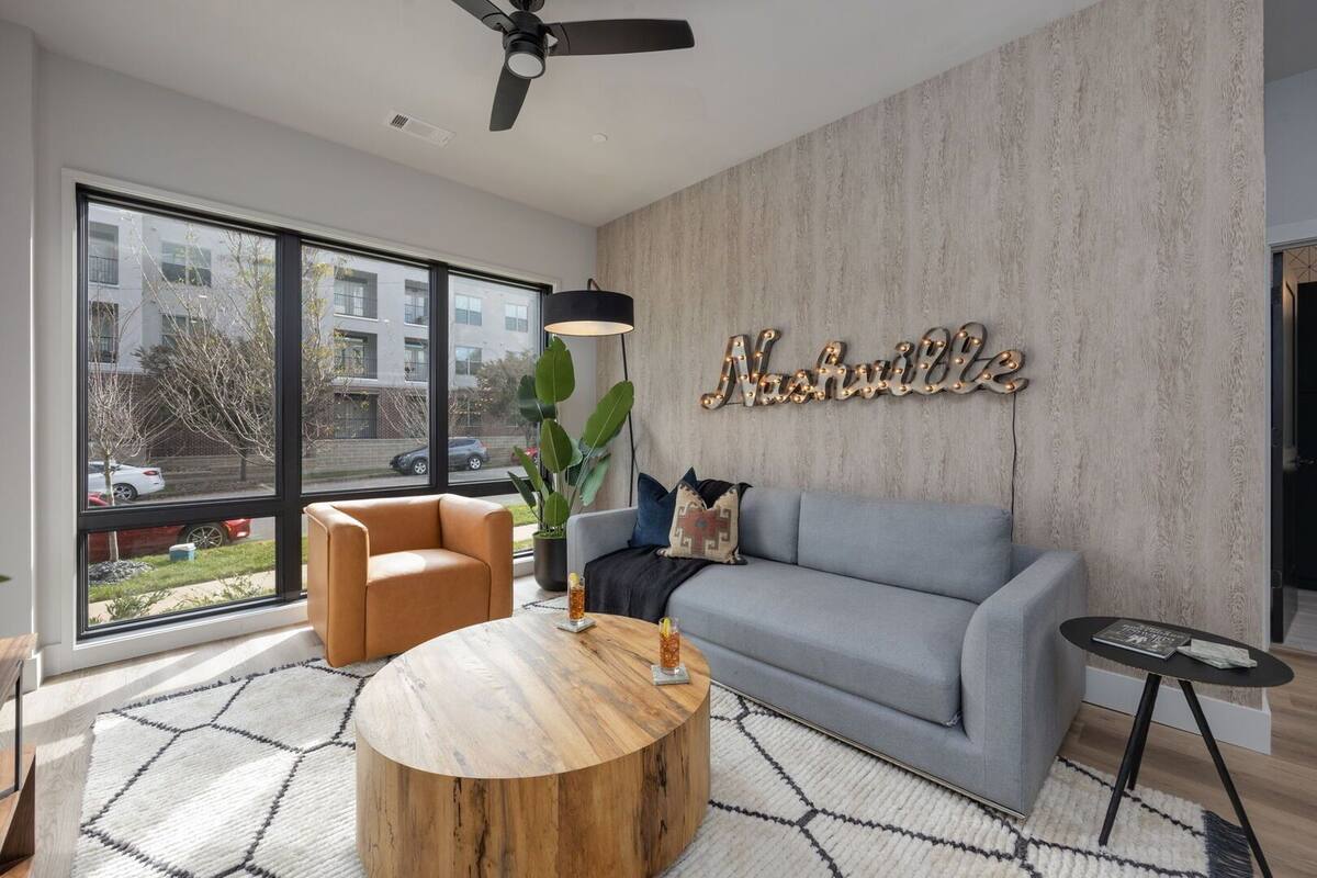 A modern living room with a large window, a grey sofa, a round wooden coffee table, and a ceiling fan.