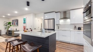 Una cocina moderna con armarios blancos, electrodomésticos de acero inoxidable, una isla central con taburetes y un comedor con una planta y obras de arte.