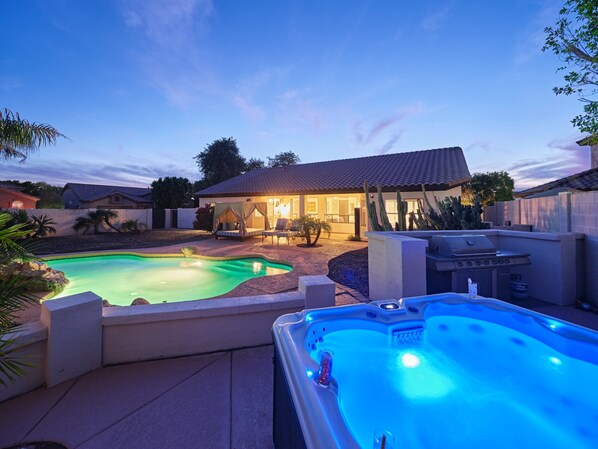A backyard with a swimming pool, a hot tub, and a covered seating area.
