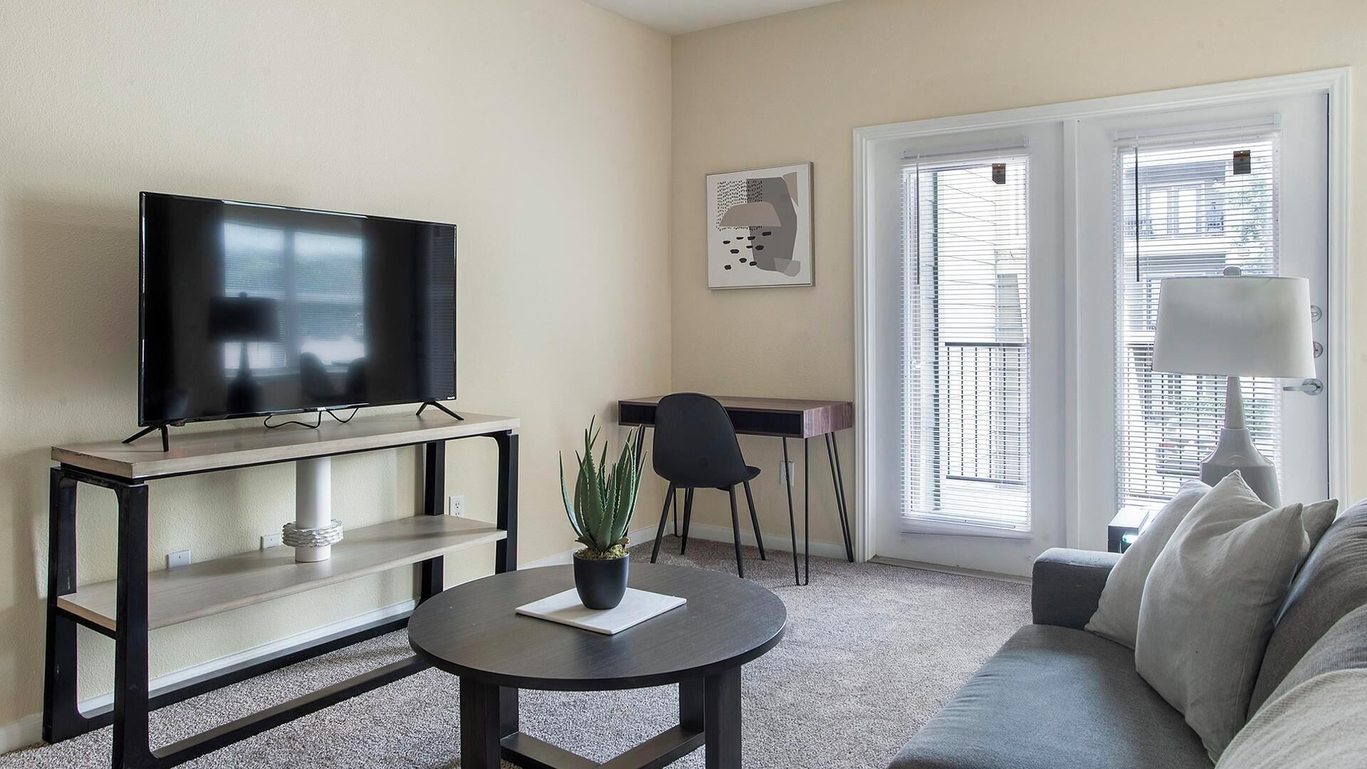 Una sala de estar con una televisión de pantalla plana sobre un mueble bajo, una mesa de centro redonda con una planta, un sofá gris, un escritorio con una silla y una ventana con persianas.