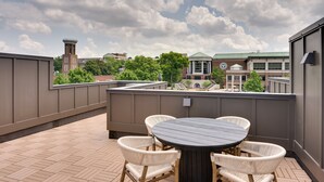 Une terrasse sur le toit, avec une table ronde et quatre chaises en osier, donnant sur un campus avec ses bâtiments et ses arbres.