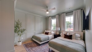 Une chambre avec deux lits, un ventilateur de plafond, une plante en pot et une fenêtre avec des rideaux.