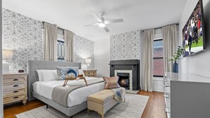 Ein Schlafzimmer mit einem Bett, Nachttischen, einem Hocker, einem Kamin, einem Deckenventilator und einem Fernseher an der Wand.