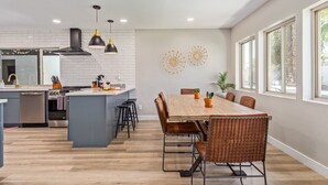 A modern kitchen with a central island, a dining area with a wooden table and wicker chairs, and a large window providing natural light. - Desert Rose-hot Tub-pool Heat Avail-fire Pit-golf! (Scottsdale)