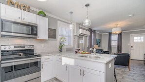 Una cocina moderna con gabinetes blancos, microondas integrado, estufa y lavamanos. Hay un comedor con una silla negra y una araña.