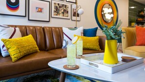 Ein modernes Wohnzimmer mit einer braunen Ledersofa, gelben Kissen, einem weißen Couchtisch mit Getränken und einer gelben Vase sowie einem farbenfrohen Wandbild.