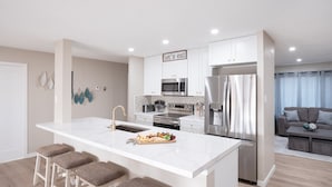 A modern kitchen with a central island, bar stools, stainless steel appliances, and a dining area with a sofa and curtains. - Blue Tides Getaway - Private Pool & Fire Pit (Scottsdale)