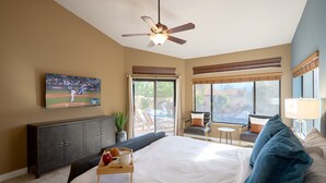 Ein Hotelzimmer mit einem großen Bett, einem Deckenventilator, einem an der Wand befestigten Fernseher, einem Esstisch mit Tablett und Blick ins Freie.