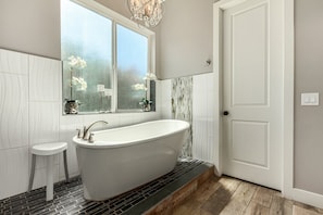 A modern bathroom with a freestanding bathtub, a small stool, a window with decorative items, and a door. - Sunny Daze in Old Town Scottsdale - Heated Pool (Scottsdale)