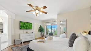Une chambre à coucher moderne avec un grand lit, un ventilateur de plafond, un téléviseur à écran plat, une table de chevet, une plante et une vue sur l’extérieur.
