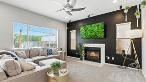 Un salon moderne avec un canapé d’angle, une cheminée, un ventilateur de plafond et une grande œuvre d’art murale aux tons verts.