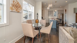 A modern kitchen with stainless steel appliances, a dining area with a glass table and cushioned chairs, and a living area with a sofa and a TV.