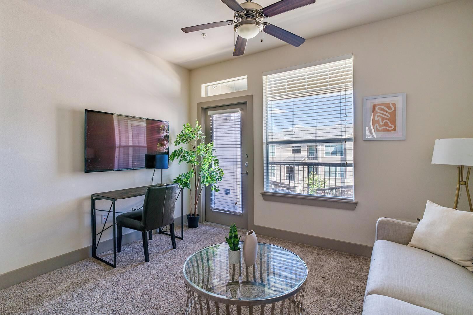A living room with a TV, a desk, a chair, a glass table, a sofa, a ceiling fan, and a window with blinds.