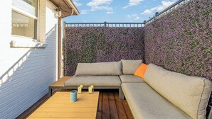 A covered outdoor seating area with a wooden table, a cushioned sofa, and a wall covered in purple flowers. - Opulent Pearl - Private Patio - Mins to Broadway (Nashville)