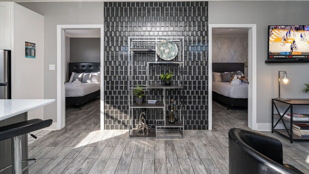 A modern living room with a grey brick-patterned wall, a central shelving unit with decorative items, a flat-screen TV, and a dining area with a white table and bar stools. - Opulent Pearl - Private Patio - Mins to Broadway (Nashville)