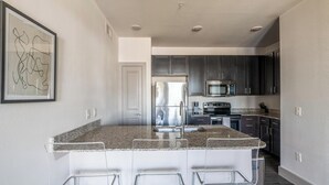 A modern kitchen with granite countertops, stainless steel appliances, and dark cabinetry. - Landing | Incredible 1BD, Pool, Gym (Houston)