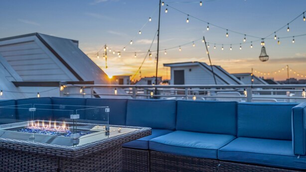 Una terraza en la azotea con una mesa de vidrio, asientos acolchados azules y luces colgantes.
