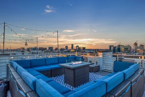 A rooftop terrace with a blue sofa set, a glass-top table with a fire feature, and string lights against a city skyline at dusk. - Nashview- Heart of Gulch - 360º Views of Downtown! (Nashville)