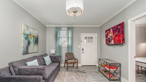 A living room with a grey sofa, a wooden chair, a coffee table, a chandelier, a painting, and a TV. - Lucent Pearl - Private Patio - Near Broadway (Nashville)