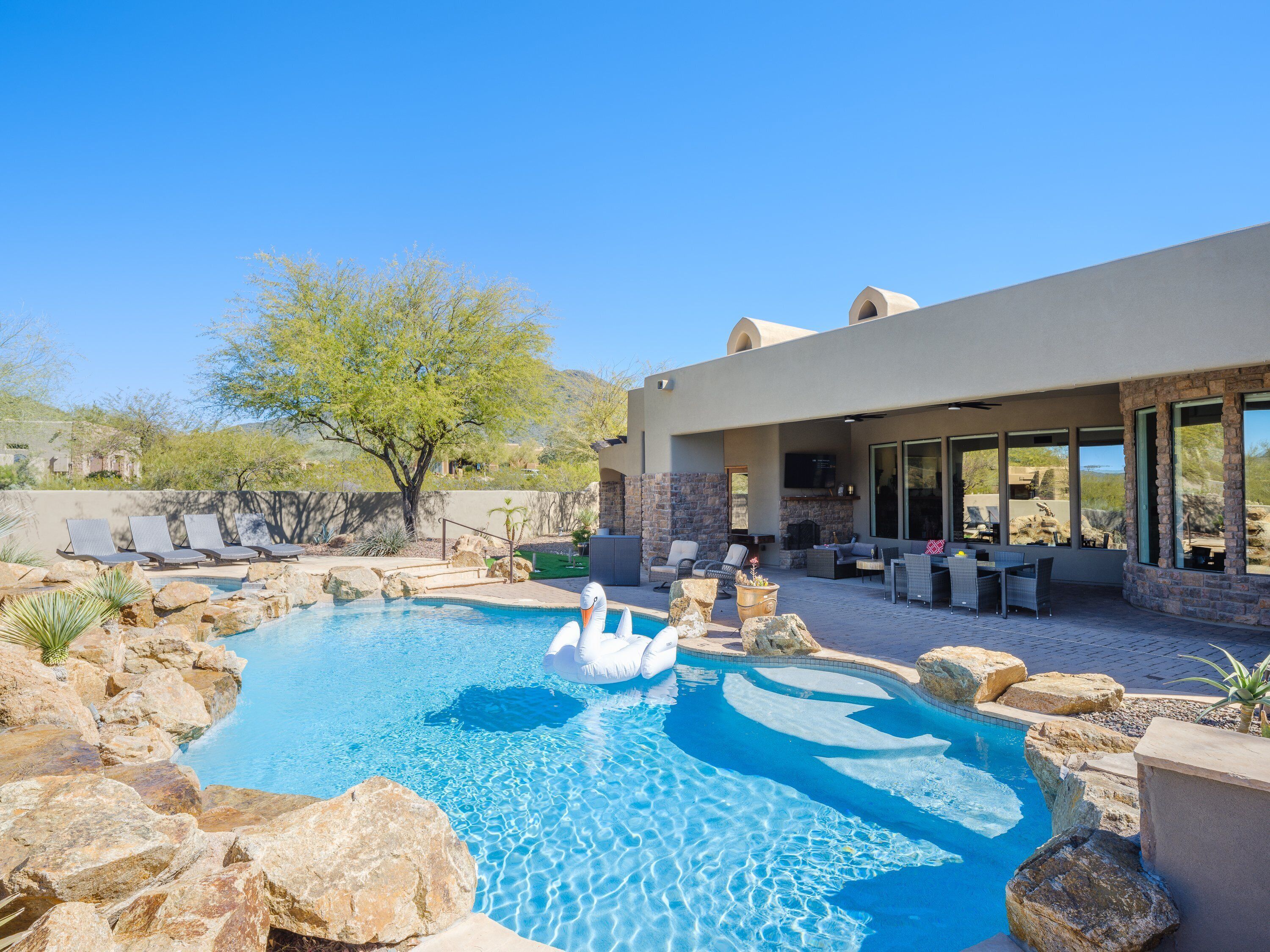 A swimming pool with rocks and lounge chairs, a stone wall, and a covered patio area with outdoor furniture.