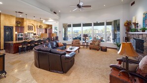 A spacious living area with a brown leather sofa, a coffee table, and a fireplace. There is a kitchen area with a bar and dining chairs. The room has large windows and a ceiling fan. - Desert Mountain Majesty - Private Spa&guest Casita (Cave Creek)