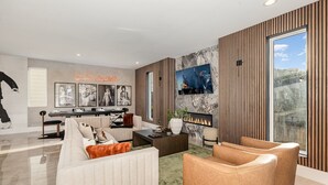 Ein modernes Wohnzimmer mit einer Couch, einem Couchtisch und Sesseln. Ein Kamin, ein an der Wand montierter Fernseher und gerahmte Bilder sind ebenfalls vorhanden.