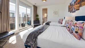 Een hotelkamer met twee bedden, een groot raam met uitzicht op een balkon, een plafondlamp en een wand met kunstwerken.