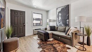 Un salon moderne avec un canapé d’angle, un pouf rond, un grand tableau abstrait, une lampe sur pied et une plante en pot.