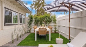 Un jardin avec une table à manger pour quatre personnes, entourée de chaises, d’un parasol à rayures et d’une petite table ronde sur laquelle se trouve une bouteille.