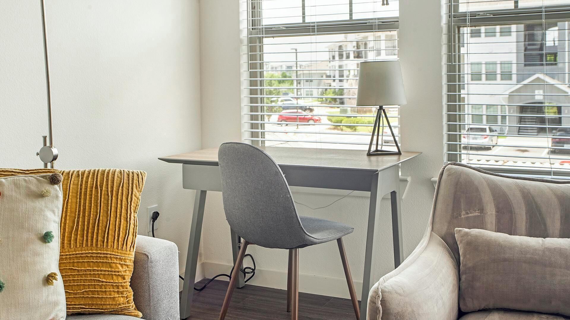Una sala de estar moderna con un sillón gris, una mesita con una lámpara y vistas a un paisaje urbano a través de amplios ventanales.