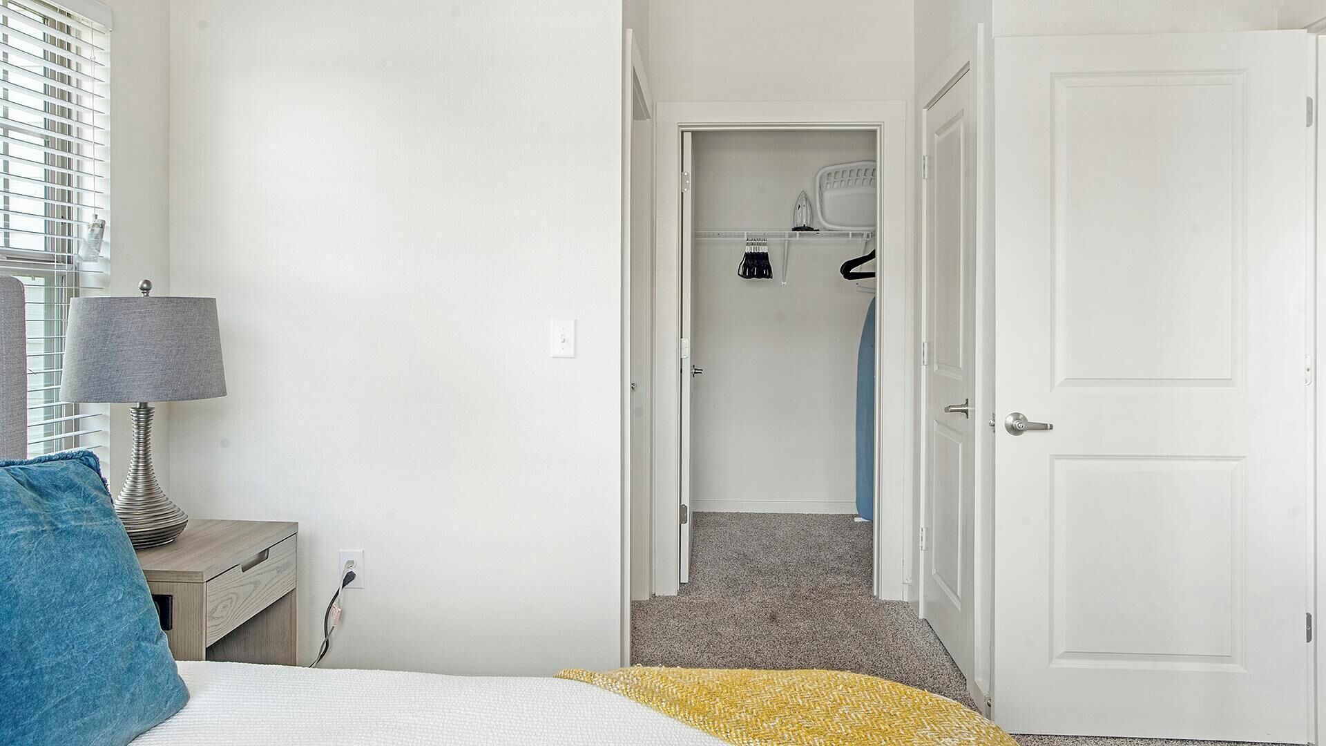 A bedroom with a bed, a bedside table with a lamp, a blue pillow, and a closet with hanging clothes.