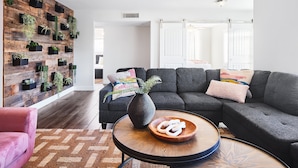 Een moderne woonkamer met een hoekbank, een houten salontafel, een wandplank voor planten en een kamerkleed met een patroon.