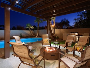 Um pátio externo com piscina, cadeiras de descanso, lareira e churrasqueira.