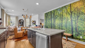 A modern kitchen with a central island, a dining area with a sofa, and a large forest mural on the wall. - Woodwind - Minutes to Broadway -walk to Breweries (Nashville)