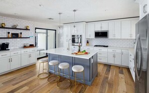 Una cocina moderna con armarios blancos, una isla central con taburetes, electrodomésticos de acero inoxidable y suelo de madera.