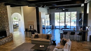 A spacious living area with a stone fireplace, a large sofa, and a coffee table. There is a kitchen area with a bar and large windows.