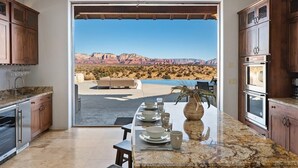 A kitchen with granite countertops, wooden cabinets, and a view of a desert landscape through an open door.