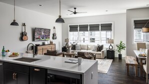 Uma cozinha moderna com uma ilha central, sala de jantar com mesa e cadeiras, sala de estar com sofá e televisão, e janelas amplas com persianas.