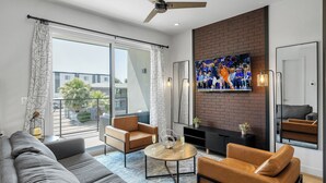 Ein modernes Wohnzimmer mit einem grauen Sofa, einem braunen Ledersessel und einem Couchtisch. Ein Fernseher ist an einer Ziegelwand befestigt, und darüber befindet sich ein Deckenventilator.