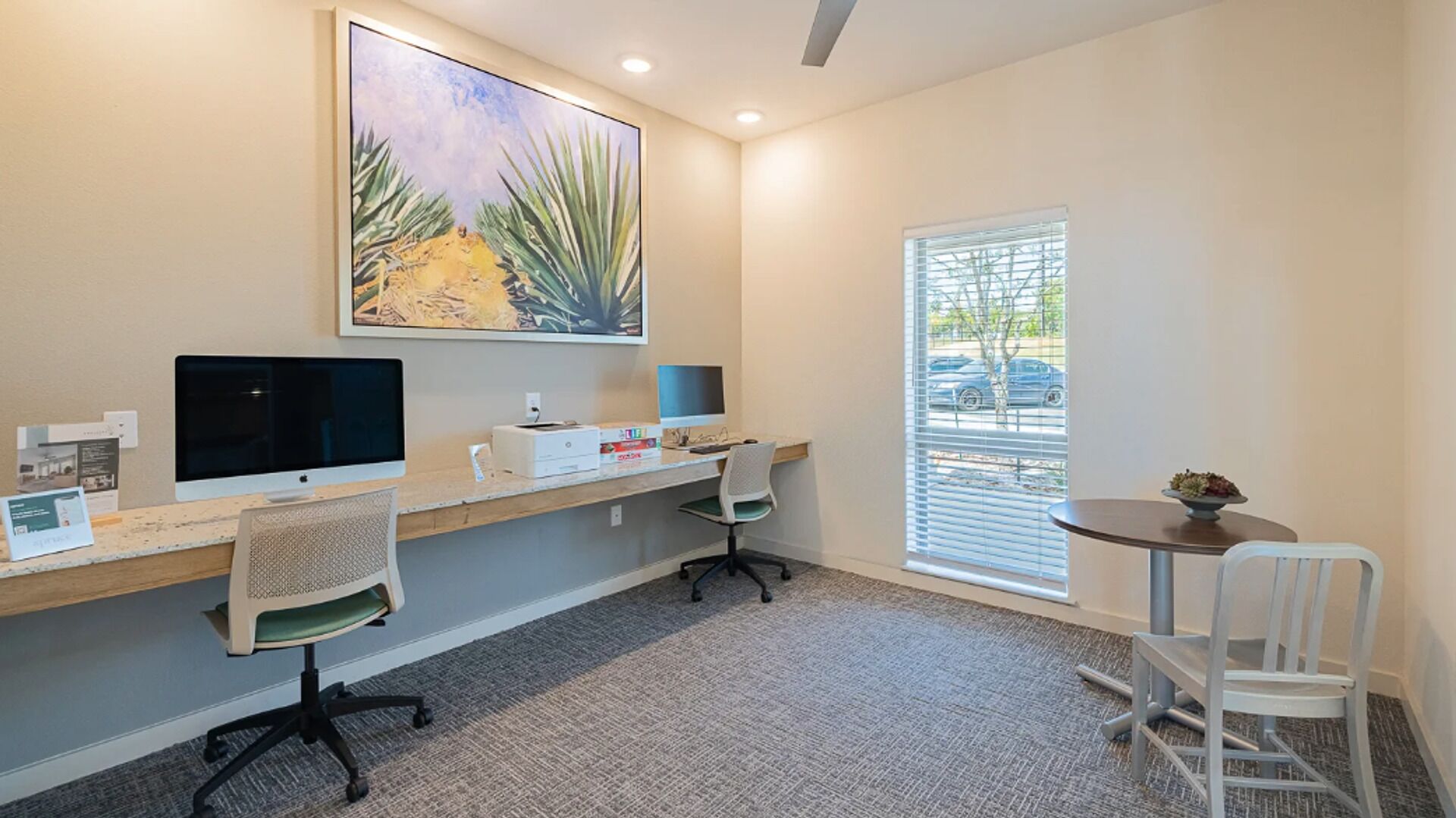 A modern office space with a desk, two monitors, a printer, and a large window with blinds.