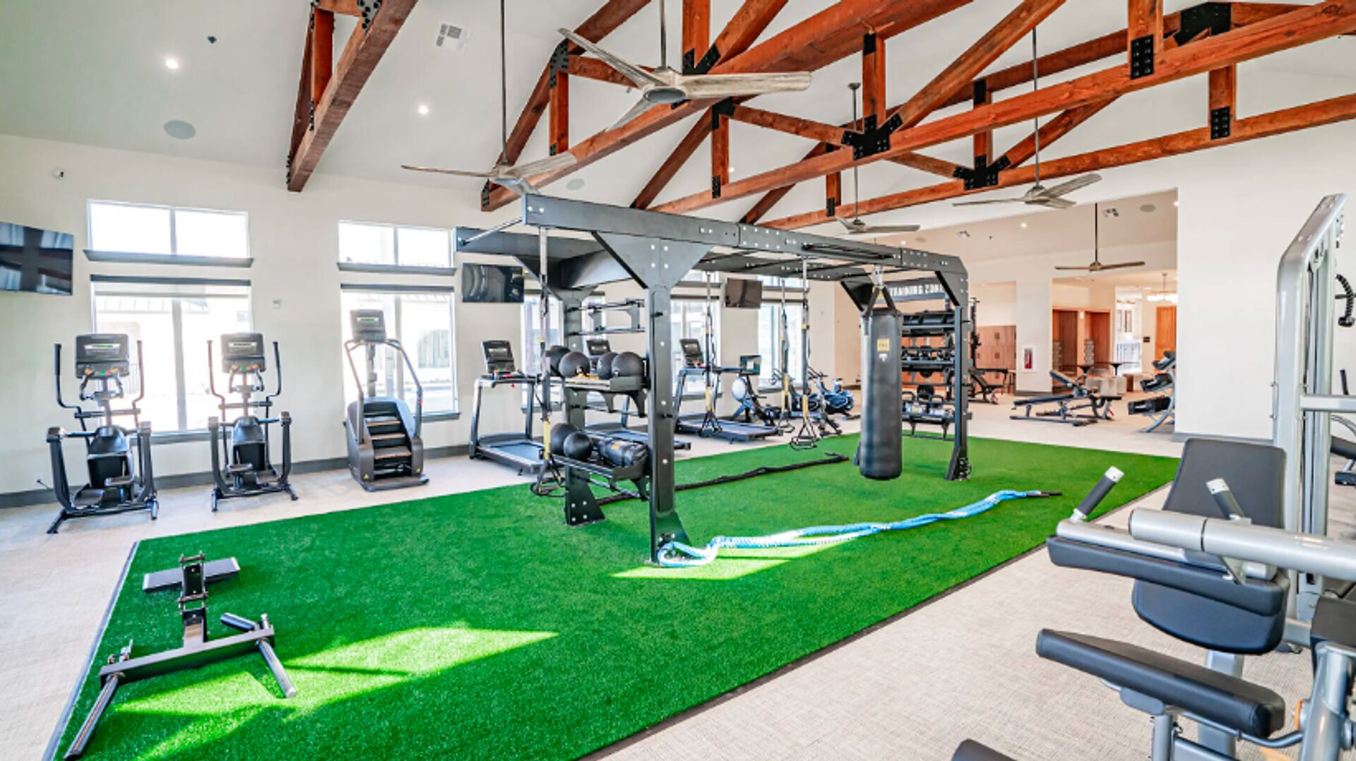 A gym with various exercise equipment, a green artificial turf area, and exposed wooden beams on the ceiling.